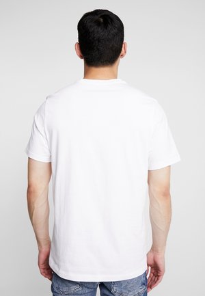 Mann mit kurzen dunklen Haaren, der ein schlichtes weißes kurzärmeliges T-Shirt und blaue Jeans trägt, von hinten zur Kamera gewandt vor schlichtem Hintergrund.