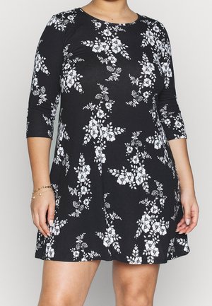 Femme portant une robe noire avec des motifs floraux blancs et des manches trois-quarts, debout devant un fond clair uni.
