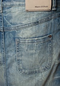Jean en denim bleu clair avec une poche arrière. Le tissu présente un motif texturé et décoloré. Une étiquette en cuir avec "Marc O'Polo" est visible.