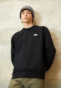 Sweatshirt negru cu guler rotund și manșete ribbate, având un mic logo alb pe piept. Asortat cu pantaloni verzi măslinii, pe un fundal neutru.