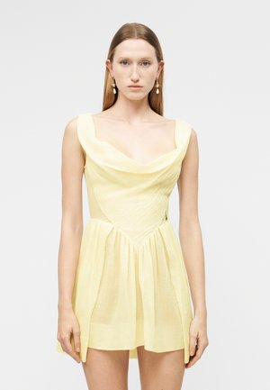 Γυναίκα με ίσια μαλλιά που φοράει αμάνικο ανοιχτό κίτρινο φόρεμα με λαιμόκοψη με πτυχώσεις και πλισέ φούστα, στέκεται μπροστά σε μια απλή φόντο.