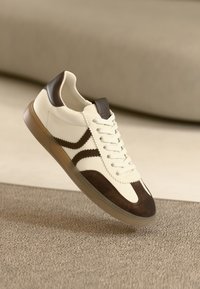 Nahast ja seemisnahast kreemikarva toss, millel on pruunid aktsendid. Omab tekstureeritud külgtriibusid, ümmargust nina ja vastupidavat kummist tallaga.