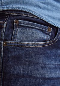 Mörkblå denimjeans med en framficka utrustad med kopparnitar och kontrasterande sömmar. Tygets struktur är något blekt på vissa ställen.