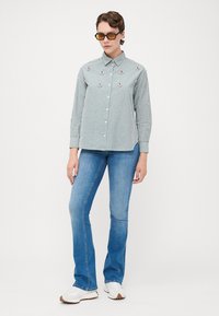 Stripete grønn og hvit skjorte med lange ermer og dekorasjoner, kombinert med lyseblå utslåtte jeans og hvite joggesko.