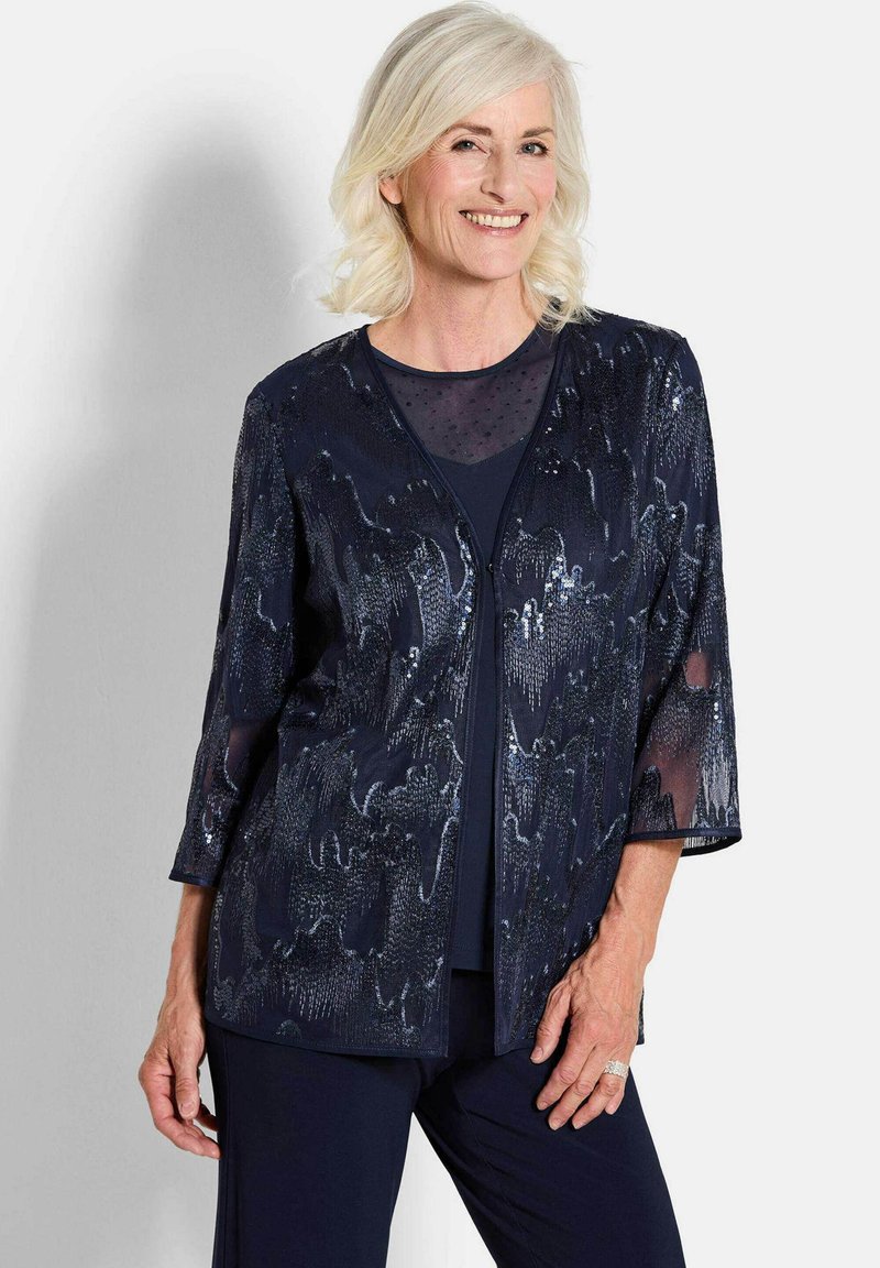 Marineblaue Jacke mit Paillettenmuster, geschichtetem Stoff, dreiviertellangen Ärmeln und offenem Design. Getragen über einem passenden Oberteil.