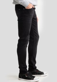 Cilvēks, kas tērpies melnās pieguļošās džinsu biksēs un melnās auduma sporta apavos ar baltu zolīti, stāv ar vienu roku kabatā pret vienkrāsainu fona sienu.