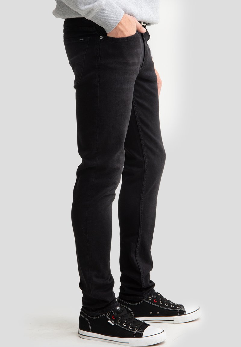 Cilvēks, kas tērpies melnās pieguļošās džinsu biksēs un melnās auduma sporta apavos ar baltu zolīti, stāv ar vienu roku kabatā pret vienkrāsainu fona sienu.