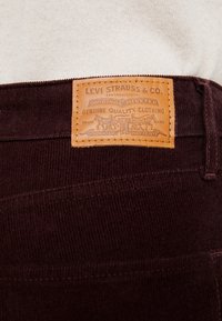 Pantalones de pana color granate con una etiqueta de piel color marrón claro que muestra "Levi Strauss & Co." y diseños intrincados; costuras y bolsillos visibles.