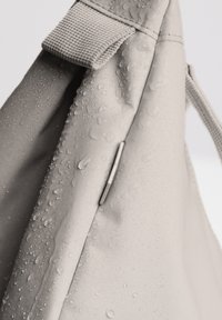 Borsa grigia resistente all'acqua con una superficie testurizzata, gocce visibili e un piccolo elemento metallico sul lato. Presenta una robusta tracolla.