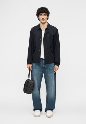 Noor mees seisab, seljas must jakk, hele särk, sinised teksad, valged tossud, hoiab parema käega väikesest musta mustriga kotist sangast.