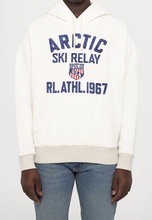 Person iført en hvid hættetrøje med blå tekst, der lyder Arctic Ski Relay Polo USA RL ATHL 1967, og lyseblå jeans på en ensfarvet baggrund.
