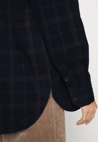 Camicia in velluto a coste blu navy con motivo a quadri rosso e verde, dotata di orlo curvo e polsini con bottone. Mano visibile che tiene la camicia.