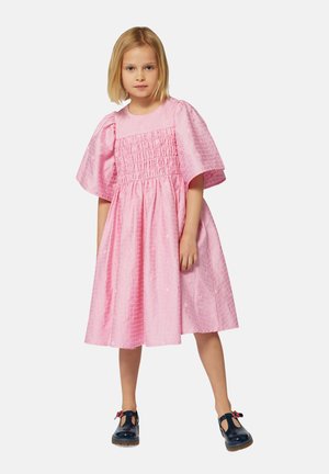 Robe rose avec un corsage smocké et de larges manches, fabriquée à partir d'un tissu texturé. Associée à des chaussures bleu marine avec un accent floral.
