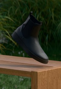 Black leather Chelsea boot with elastic side panels, minimalist design, rounded toe, and a flat rubber sole, displayed on a wooden surface.