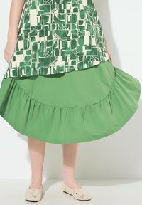 Vestido verde de capas con un top estampado que presenta diseños geométricos en verde oscuro y crema. Abajo, un dobladillo azul sólido con volantes. Zapatos de ganchillo beige.