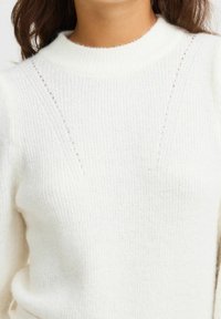 Pull en tricot blanc avec une texture côtelée, un col montant et des détails de couture décoratifs le long des épaules. Matière douce et confortable.