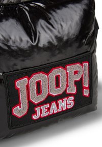 Czarna, błyszcząca tkanina puchowa z prostokątną naszywką. Naszywka zawiera napis "JOOP!" w wypukłym szarym i czerwonym haftem na czarnym tle.