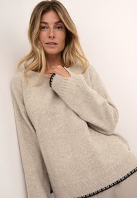 Beżowy sweter dzianinowy o luźnym kroju, z czarnymi ściągaczami przy mankietach i na dole. Miękka tekstura, szeroki dekolt i widoczne szwy dodają charakteru.