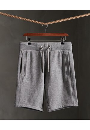 Grå shorts med snøre og sidelommer hængende på en træbøjle, som er klemmet fast til et reb mod en mørk baggrund.