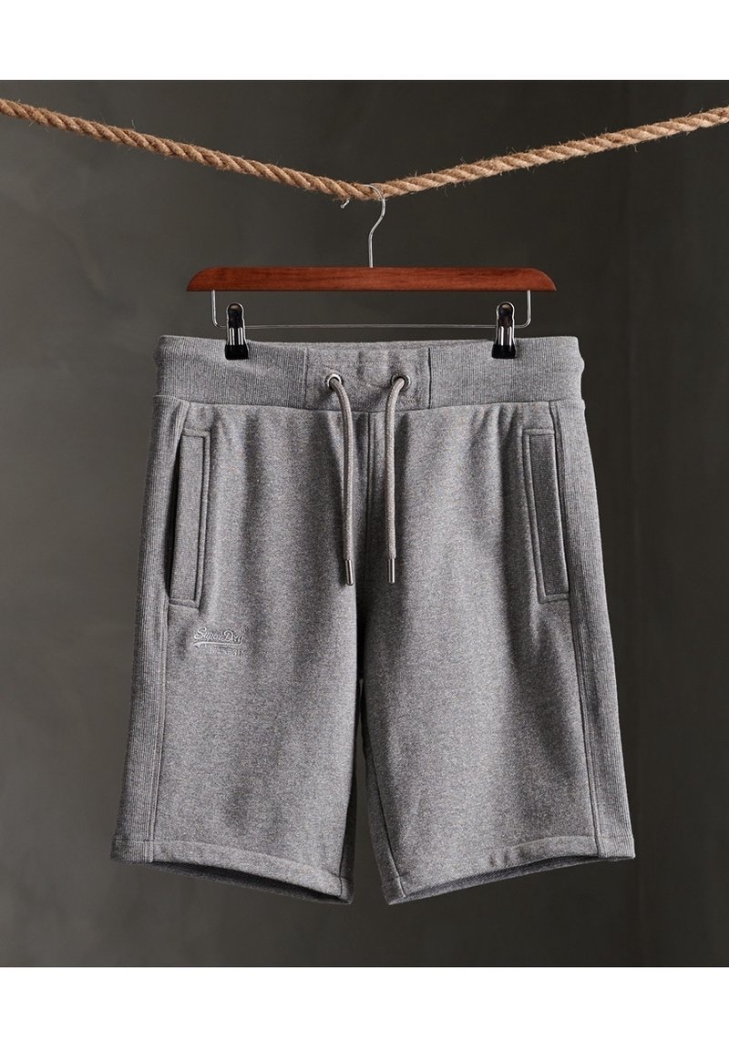 Short gris à cordon avec poches latérales suspendu à un cintre en bois accroché à une corde sur un fond sombre.
