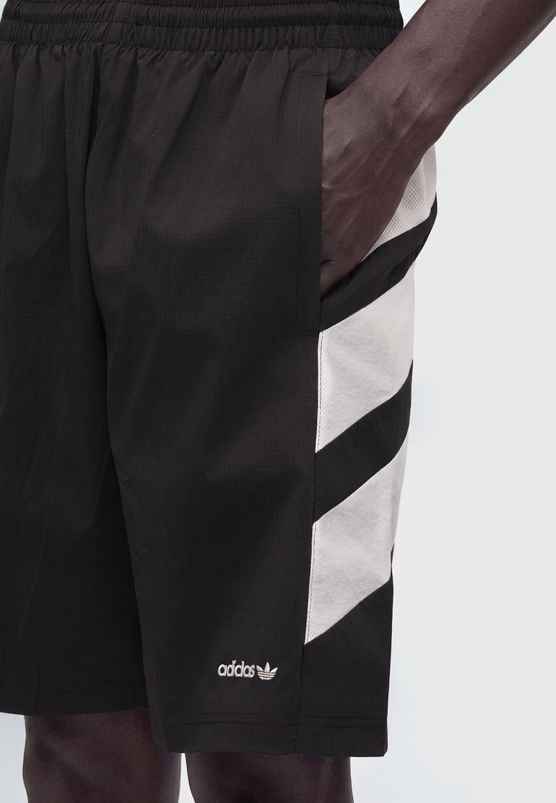 Sorte Adidas shorts med hvide diagonale striber på siden og en hånd placeret inde i den højre lomme.