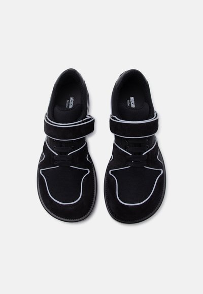 Μαύρα sneakers χωρίς κορδόνια με λευκή επένδυση, ρυθμιζόμενους ιμάντες με βέλκρο, στρογγυλεμένες μύτες και πάνελ από ύφασμα mesh, φαίνονται από πάνω σε λευκό φόντο.