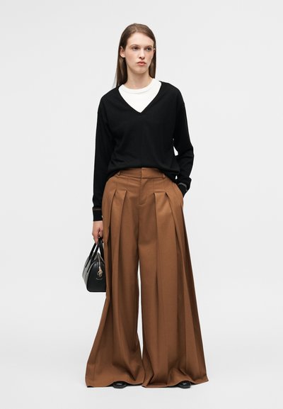 Μαύρο πουλόβερ με V-λαιμόκοψη συνδυασμένο με καφέ παντελόνι ψηλόμεσο και με πιέτες. Το μοντέλο κρατά μια μικρή μαύρη τσάντα χειρός. Μινιμαλιστικός σχεδιασμός με ομαλές υφές.