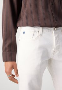 Des jeans en denim blanc présentent une coupe slim avec une fermeture à bouton, un matériel en laiton et un petit logo bleu sur la poche avant. Le modèle porte une chemise à rayures brunes.