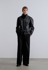 Fekete bőrdzseki álló gallérral, cipzáras záródással és oldalzsebekkel, lazán álló fekete nadrággal és fényes fekete cipőkkel párosítva.