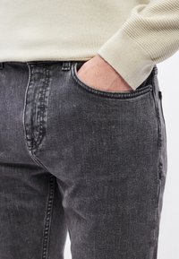 Μαύρο τζιν από τζιν με ξεθωριασμένο φινίρισμα, με μπροστινή τσέπη και εφαρμοστή σχεδίαση. Συνδυάζεται με ανοιχτό μπεζ πουλόβερ.