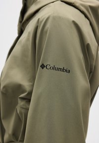 Gros plan sur la manche d'une veste vert olive avec le logo noir de Columbia brodé près de l'épaule.
