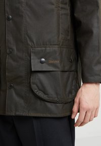 Waxed cotton jacket in dark olive green with snap buttons, a front pocket, and "Barbour" logo in brown embroidery on the pocket.