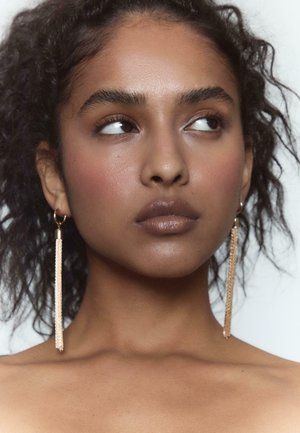 Lange, guldfarvede fringtørhæng har hængende fra hoops. Modellen har en glat teint, mørkt bølget hår og diskret makeup.