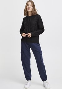 Svart stickad tröja med strukturerade kabelmönster, i kombination med marinblå joggerbyxor med sidofickor och elastiska muddar, avslutat med vita sneakers.