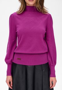 Kobieta w fioletowym swetrze z długim rękawem i golfem, z bufiastymi rękawami, oraz czarnej skórzanej spódnicy, stojąca na tle jednolitym.