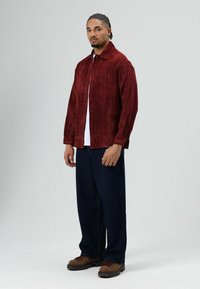 Rode ribfluwelen overhemd met knopen, witte T-shirt eronder, donkerblauwe wijde broekspijpen en bruine veterlaarzen met zwarte zolen.