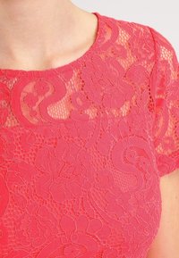 Korálový krajkový top s květinovými vzory, průhlednou strukturou a krátkými rukávy. Zaoblený výstřih dodává designu něžnost.