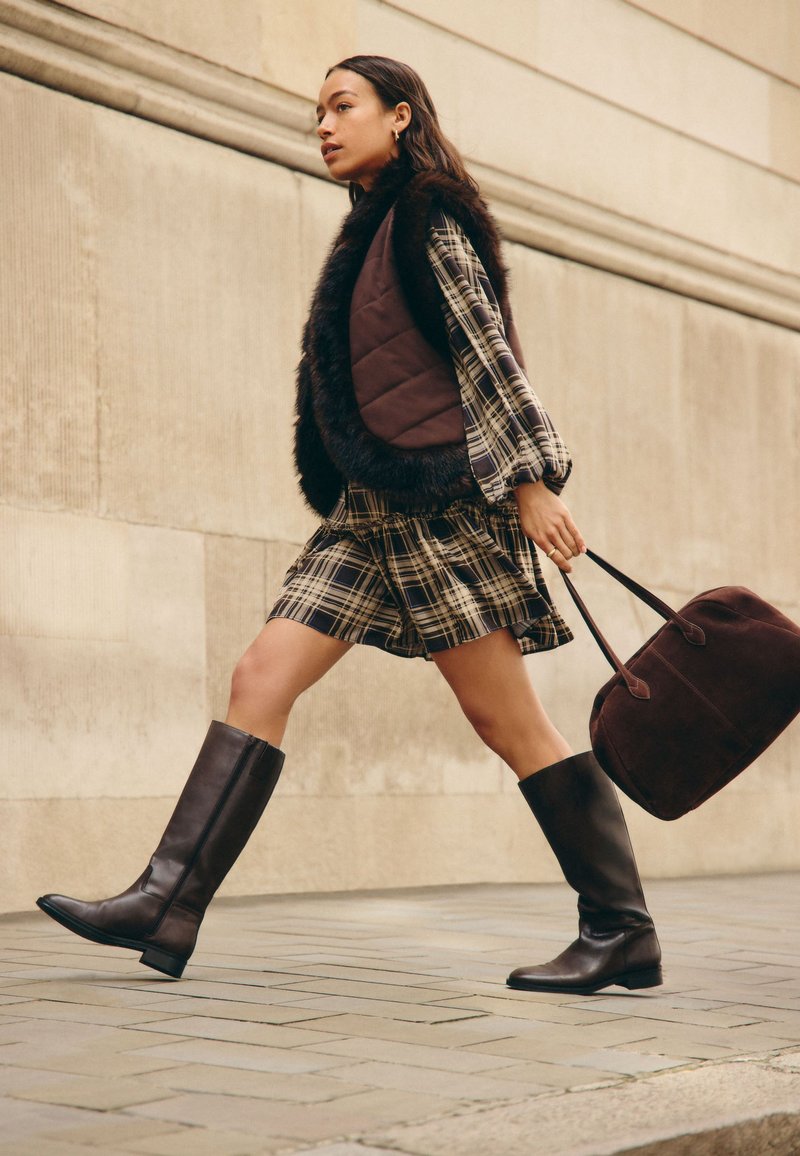 Donna che cammina sul marciapiede della città indossando un vestito a quadri con gilet bordato di pelliccia, stivali al ginocchio e porta una grande borsa scura.