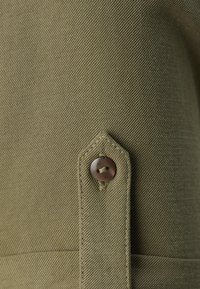 Olivengrøn stof med en struktureret finish, med en spids flap og en brun knapdetalje. Designet viser syede detaljer langs kanterne.