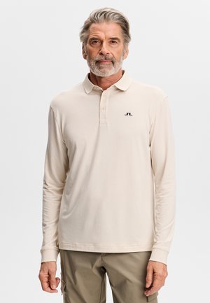 Homme d'âge moyen aux cheveux gris et à la barbe, portant un polo beige à manches longues et un pantalon kaki, se tenant devant un fond uni.