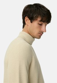 Beiger Strickpullover mit hohem, geripptem Kragen und strukturiertem Muster, von der Seite gezeigt mit glatter Oberfläche und enganliegender Silhouette.