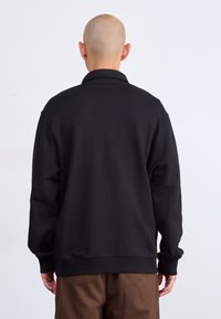 Maglione nero con colletto alto e vestibilità ampia, con maniche lunghe e polsini a coste, indossato sopra pantaloni marroni.