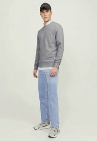 Young man standing, wearing a black cap, grey sweatshirt over a striped shirt, light blue jeans, and grey sneakers, against a plain light background.