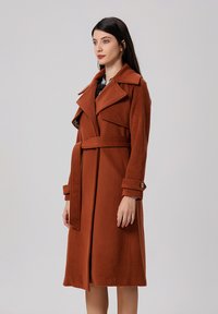 Manteau en mélange de laine marron avec une taille ceinturée, des revers larges et des poignets boutonnés. Présente une texture lisse et un design longueur genou.