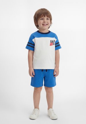 Νεαρό αγόρι στέκεται και χαμογελά φορώντας μπλε και άσπρο t-shirt GAP, μπλε σορτς και λευκά αθλητικά σε λευκό φόντο.