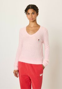 Pull en V à col plongeant, en rose clair et pelucheux, avec des manches longues et un ourlet côtelé, arborant un petit écusson multicolore sur la poitrine.