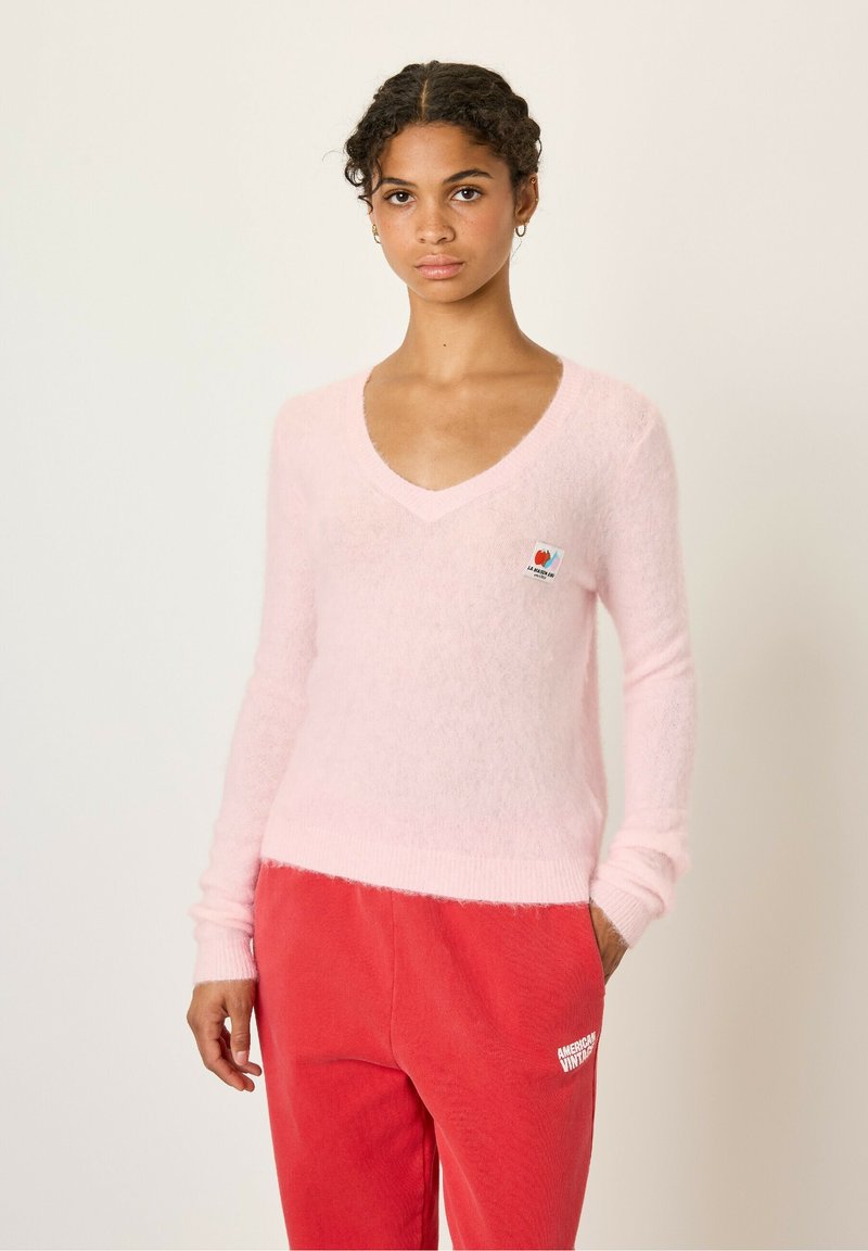 Pull en V à col plongeant, en rose clair et pelucheux, avec des manches longues et un ourlet côtelé, arborant un petit écusson multicolore sur la poitrine.