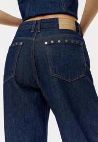 Dark wash denim jeans with a plain waistband, brown leather patch, and metal studs on back pockets. Features traditional stitching.