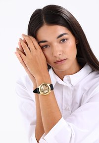 Montre dorée avec un cadran rond, bracelet en silicone noir et détails décoratifs sur le cadran. Le modèle porte une chemise blanche à boutons.