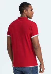 Röd polo skjorta med krage, korta ärmar och vit kant runt ärmsluten och kragen. Tyget verkar slätt med en figursydd design.